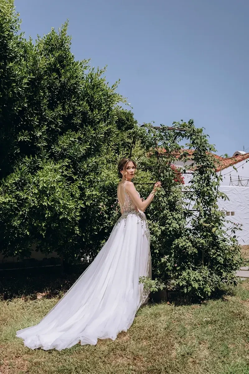 Bride in a flowing white gown stands in a garden, touching a leafy arch. Sunlit setting with clear blue sky creates a serene, elegant ambiance.