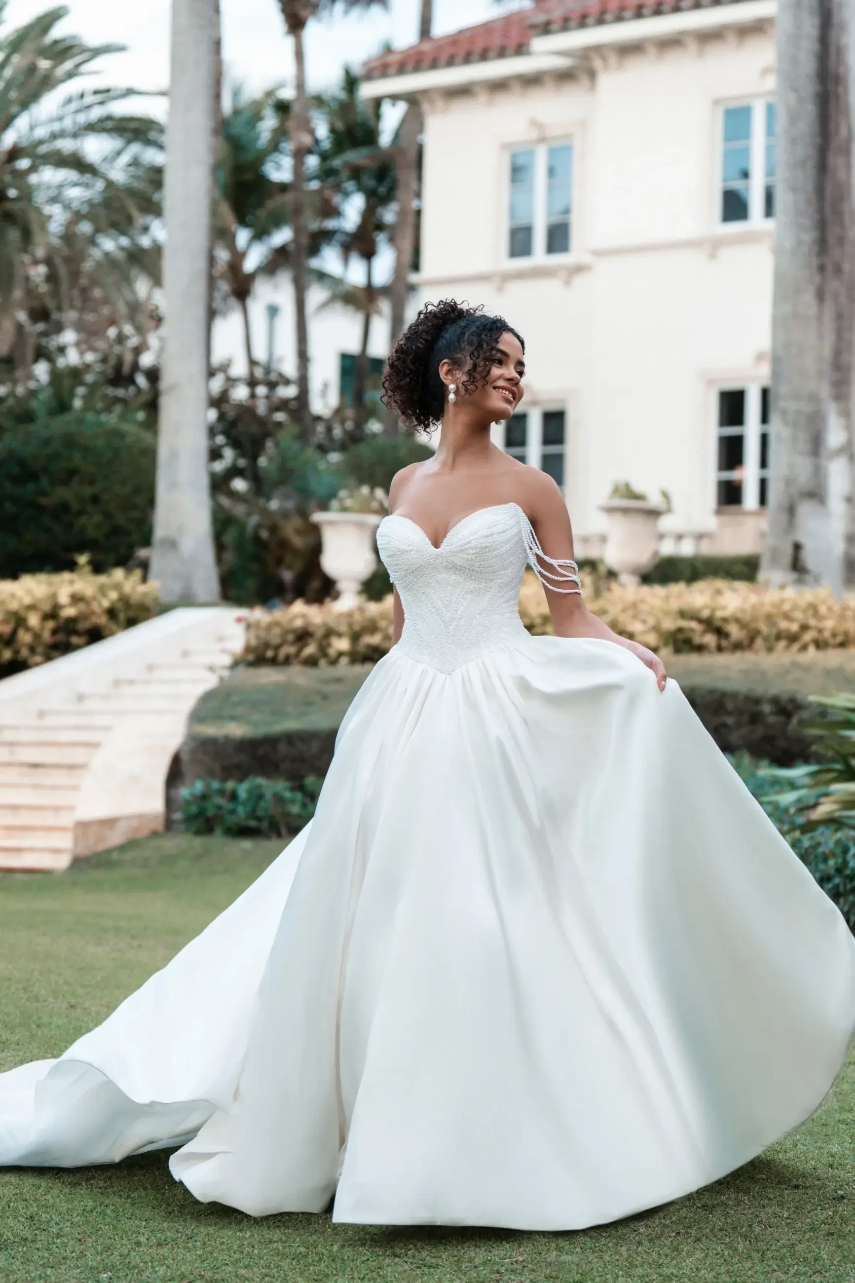 A bride in a flowing white gown with lace details stands joyfully on a lawn. Behind her is a grand villa with lush palm trees and hedges.