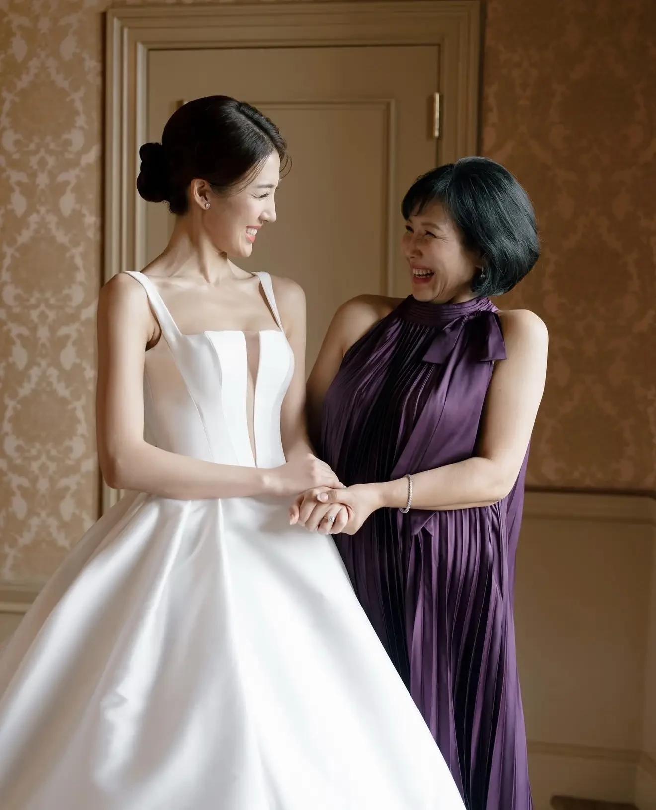 A bride in a white gown smiles warmly at a woman in a purple dress. They stand indoors, hand in hand, conveying joy and a loving connection.