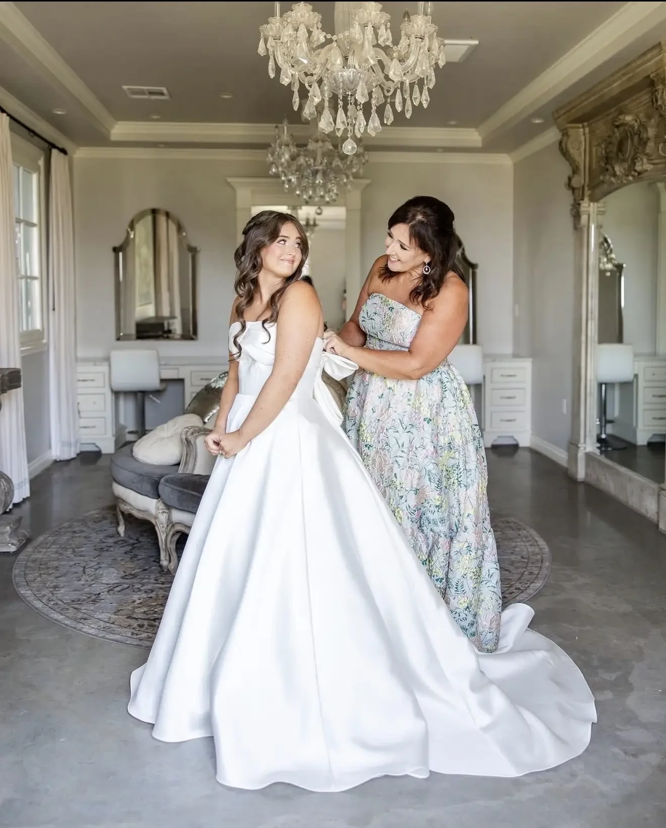 A bride in a white gown stands in an elegant room while a woman in a floral dress adjusts her dress. The mood is joyful and intimate, under a chandelier.