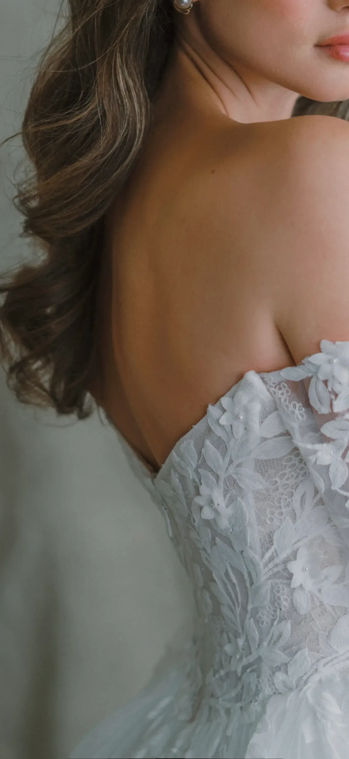 A close-up of a woman's shoulder and the back of a white lace dress, with soft waves of hair cascading down her back.