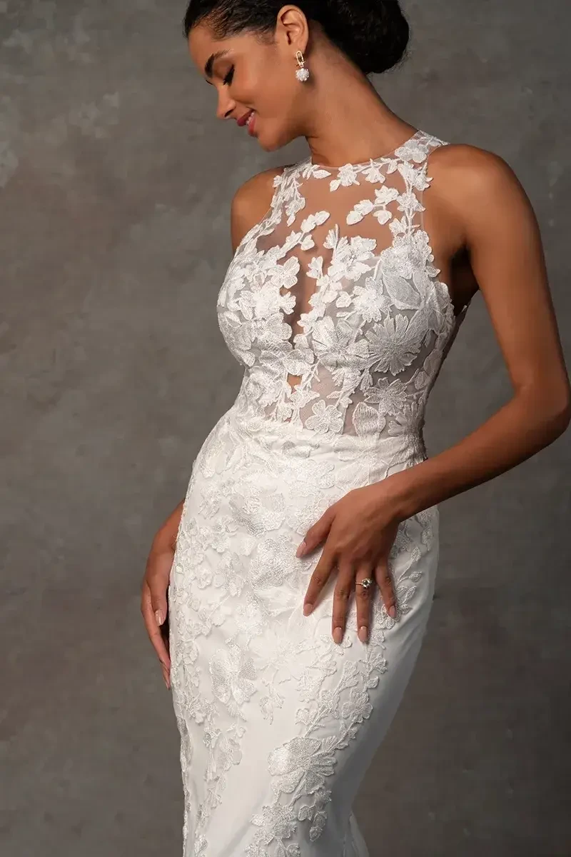 A woman in an elegant white lace wedding dress poses against a gray background. The dress features intricate floral patterns and a sheer neckline. She looks down with a serene smile.
