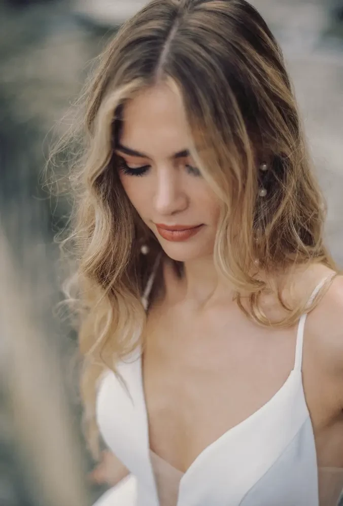 A woman with wavy hair wearing a white dress, looking down thoughtfully.