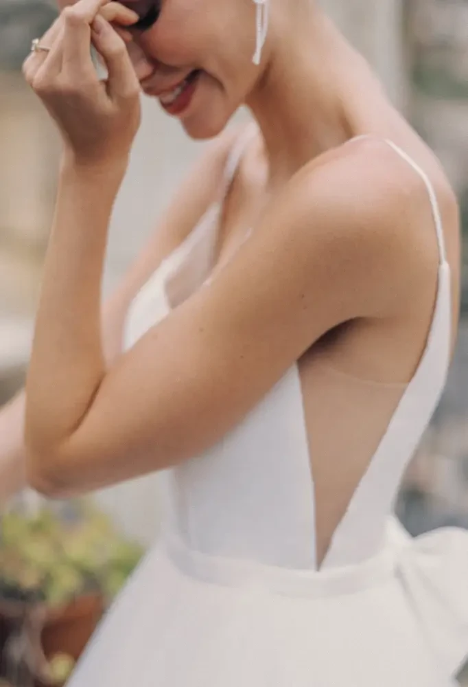 A woman in a white dress with a low back, smiling while touching her face.
