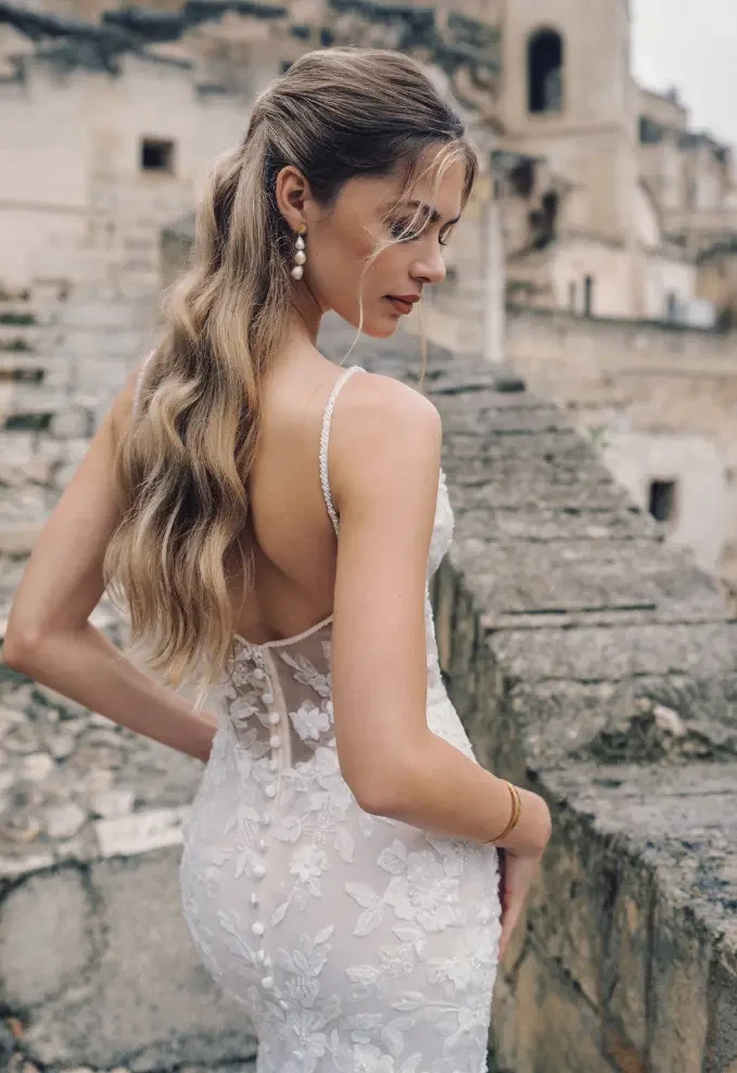 A woman in a backless wedding dress with intricate floral details poses outdoors, showcasing her long wavy hair and natural makeup against a rustic stone backdrop.