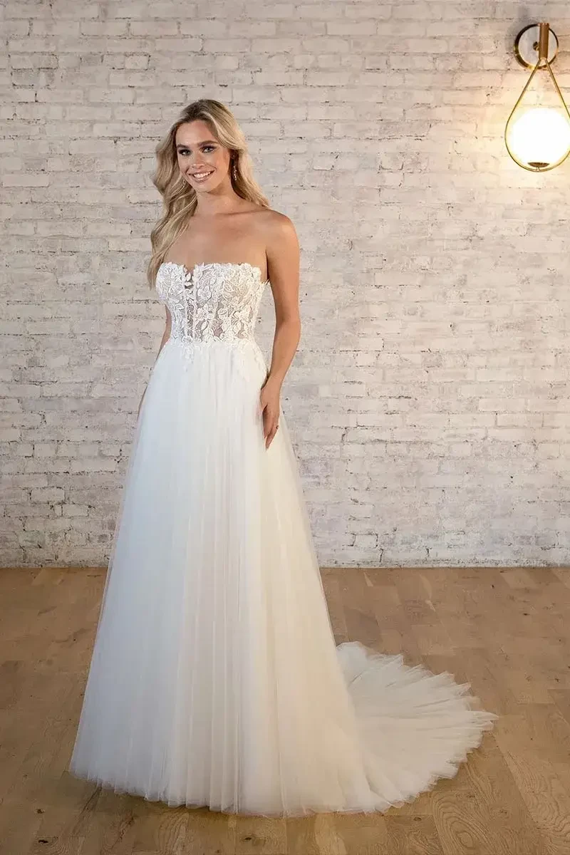 A woman in a strapless lace and tulle wedding gown stands on wooden flooring, smiling against a textured white brick wall with a glowing light fixture.