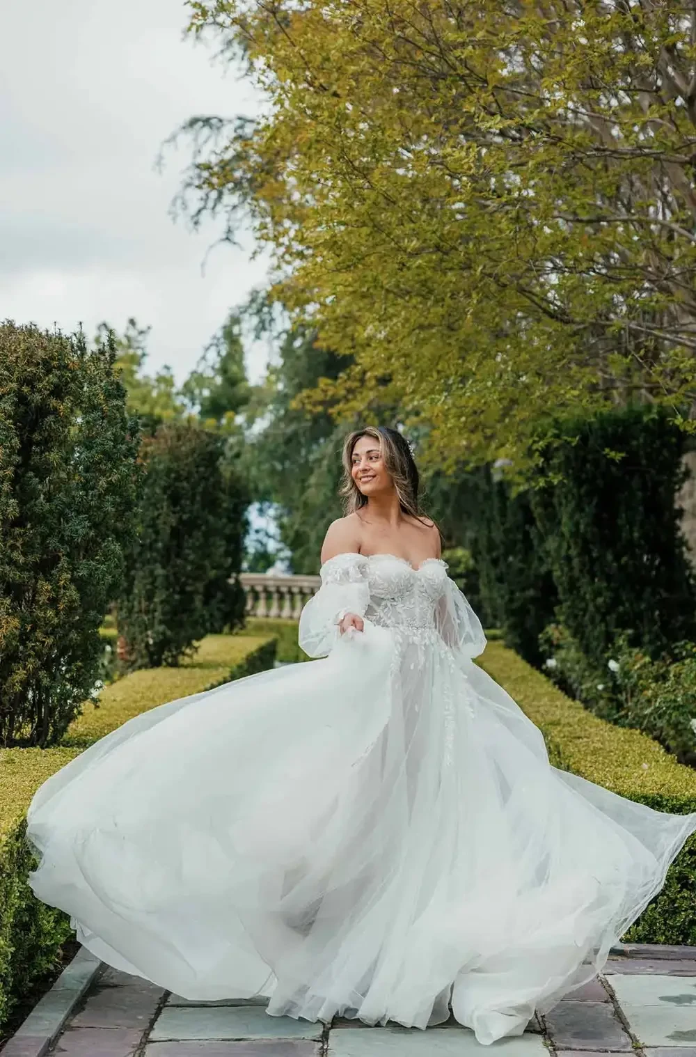 A woman in a flowing white dress twirls in a garden, surrounded by greenery and trees.