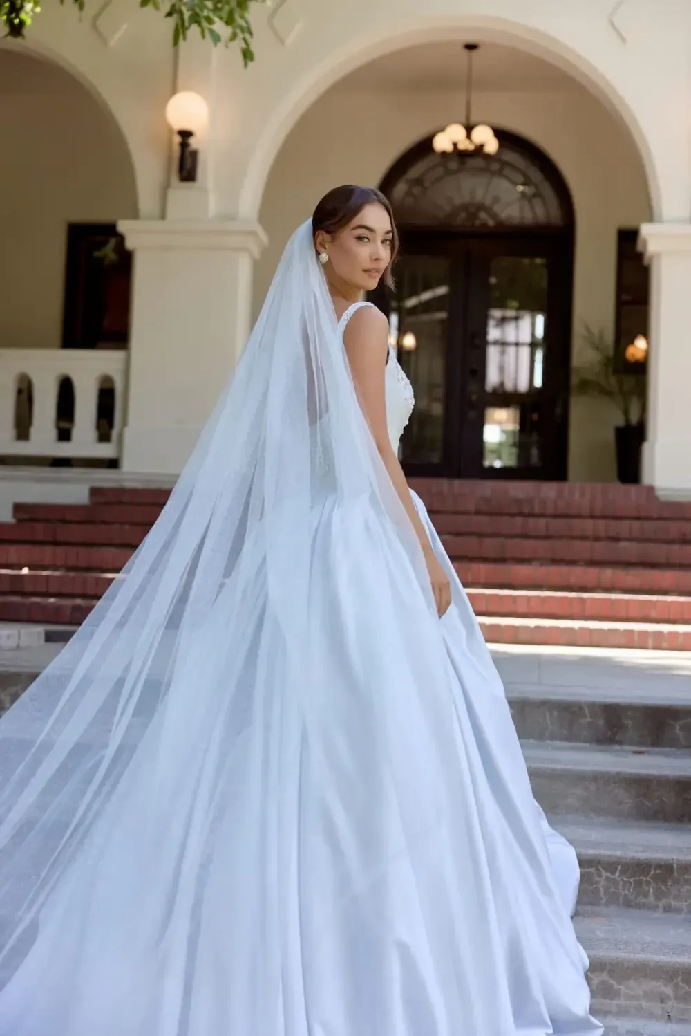 A bride in a flowing white gown and veil stands gracefully on steps, looking back over her shoulder.