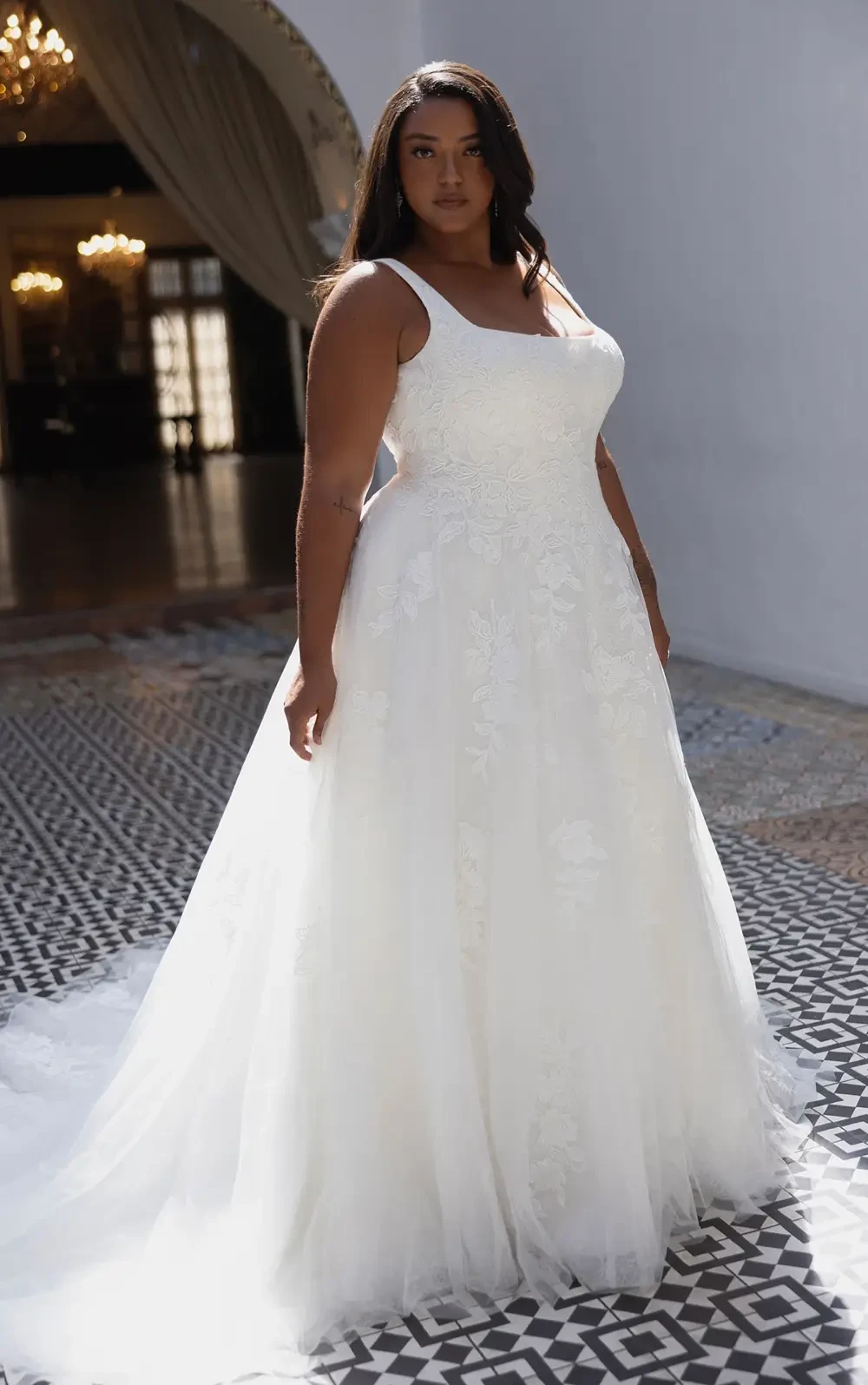 A woman in a white wedding dress stands elegantly, with a full skirt and floral details, in a grand indoor setting.