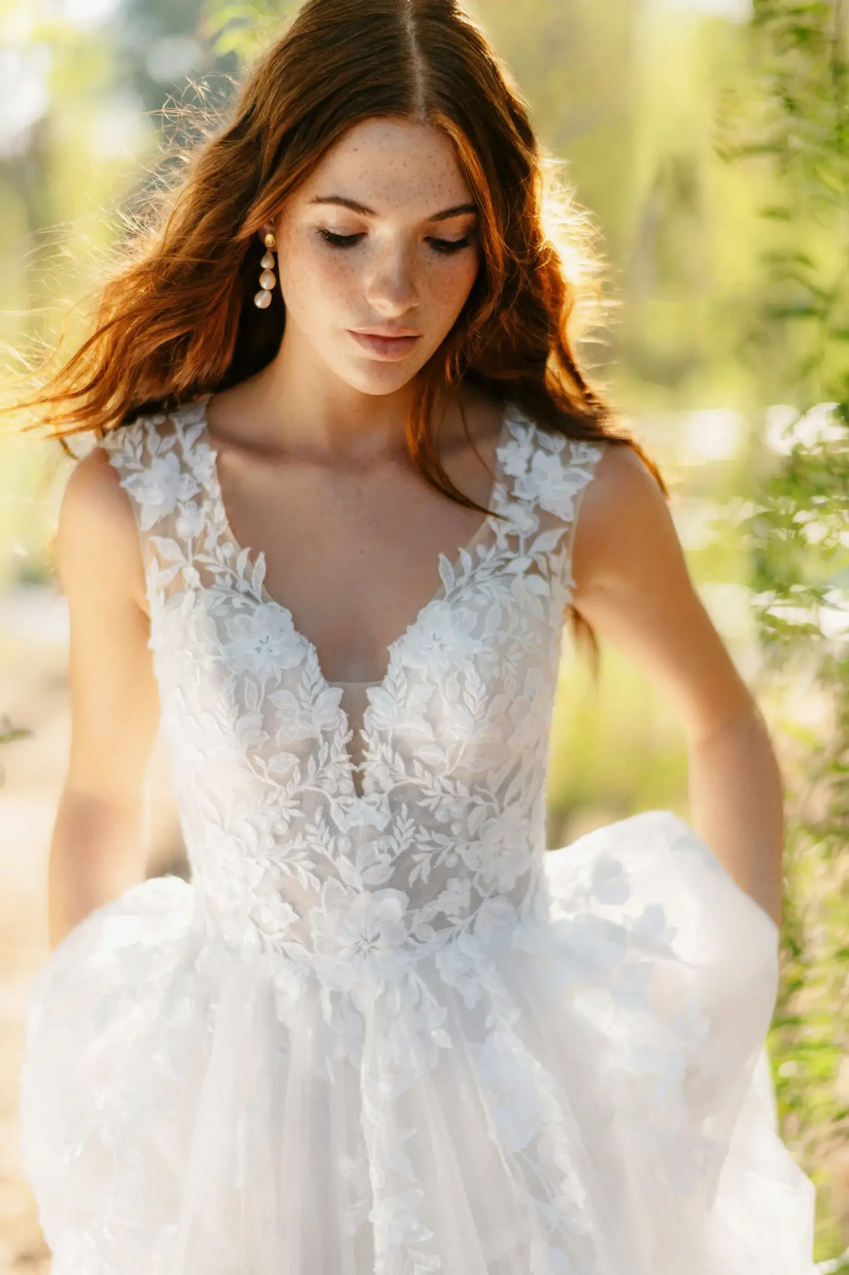 A woman wearing a white floral wedding dress, standing in a natural setting with soft sunlight filtering through. She has long, flowing hair and is looking downwards.