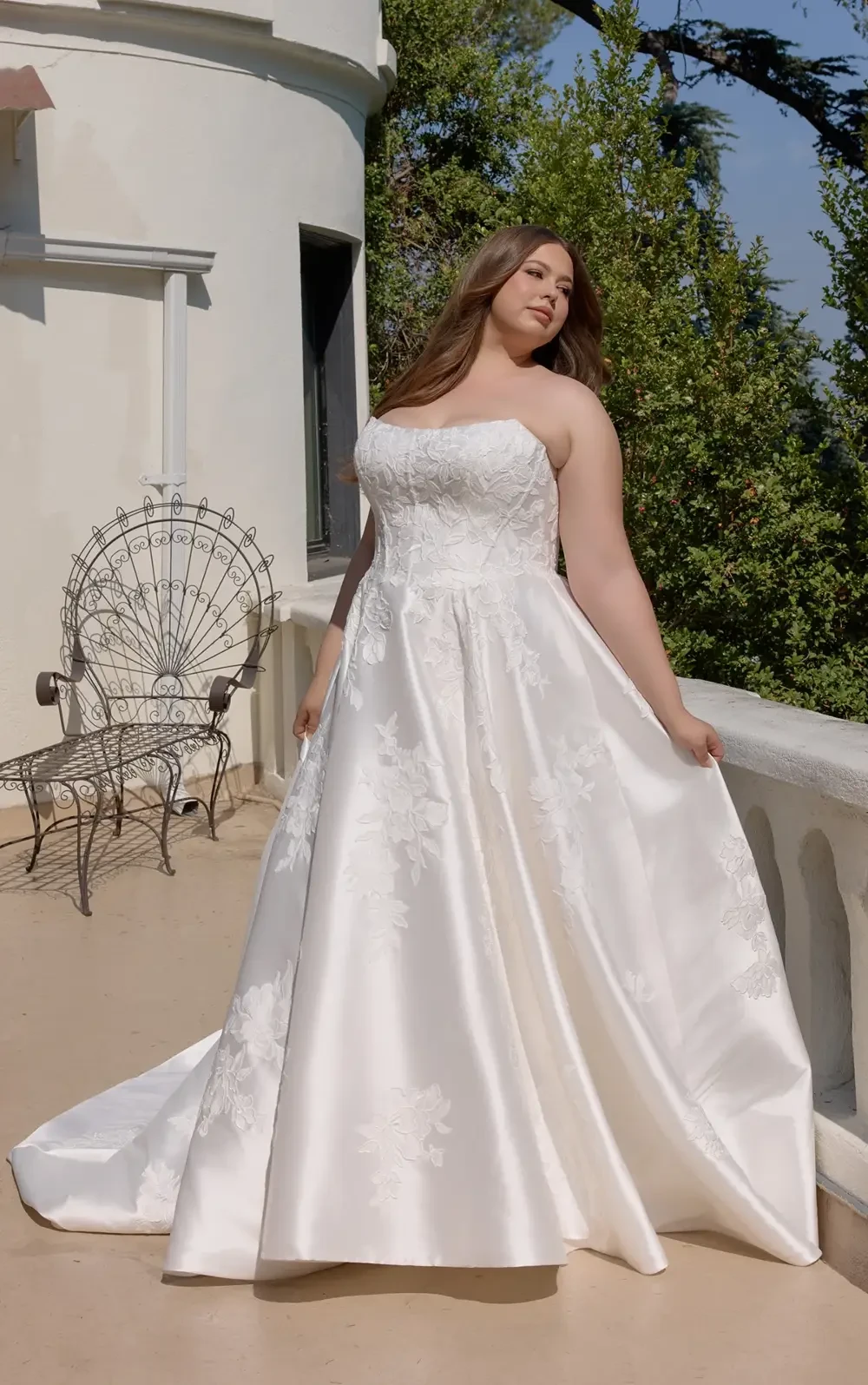 A plus-size woman in a flowing white wedding gown stands on a balcony surrounded by greenery.